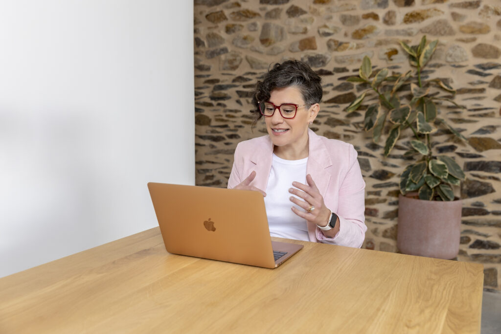 Hannah is sitting at her laptop. She is speaking to a client on a call, and is using her hands expressively. She is wearing a light pink jacket.